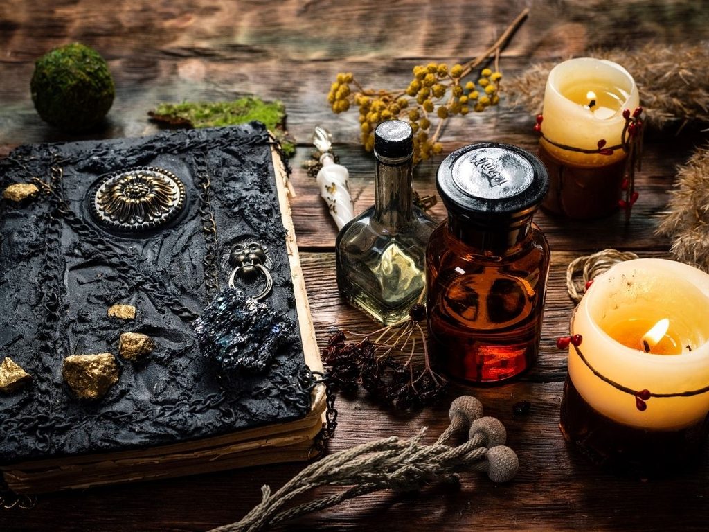 Antique-style grimoire beside glass jars, dried herbs, and lit candles on a rustic wooden table. 